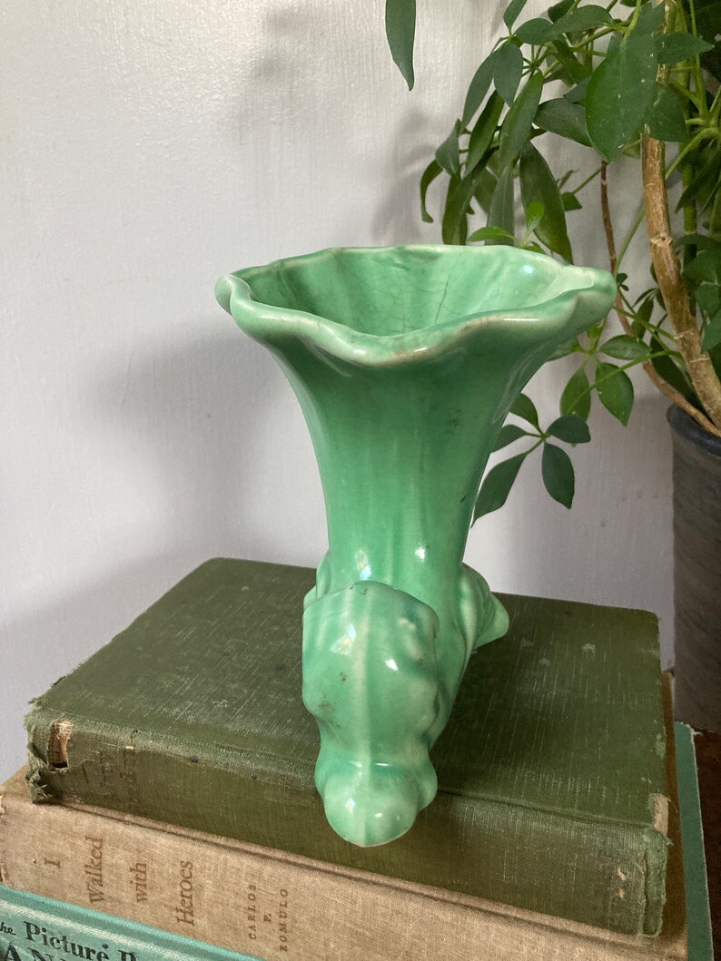 May include: A green ceramic vase with a trumpet-shaped opening and a sculpted base. The vase is sitting on a stack of books.