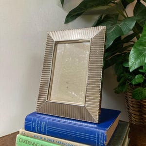 May include: A rectangular silver-toned picture frame with a vertical ribbed texture on the border. The frame is positioned upright on top of two stacked hardcover books, one blue and one green, with visible text on their spines.