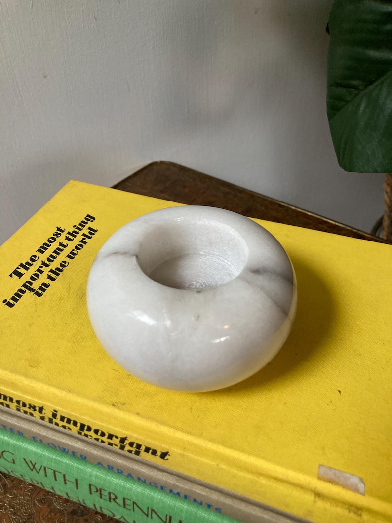May include: White marble candle holder with a round shape and a smooth, polished surface. The holder has a small, circular opening in the center.