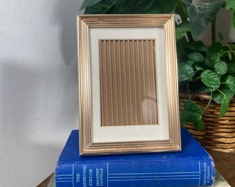May include: A rectangular gold-colored picture frame with a cream-colored mat, resting on a stack of books. The books are blue, beige, and green. The frame is empty, with a corrugated cardboard backing.
