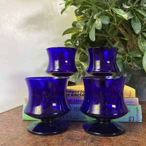 May include: Four cobalt blue glass cups with a flared base and a rounded top. The cups are arranged in a pyramid shape on a wooden surface.