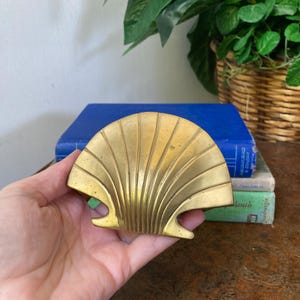 May include: A brass seashell-shaped object with a fan-like design. The object is held in a hand, and the background includes books and greenery. The seashell has a golden hue and a textured surface, suggesting it is made of metal.