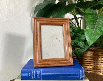 May include: A rectangular wooden picture frame with a light brown finish, resting on a stack of books. The books are blue, green, and off-white, with titles visible. Lush green plants are in the background.