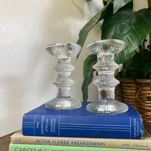 May include: Two clear, textured glass candle holders with a pedestal base and a flared top. The candle holders are displayed on top of a stack of vintage books with blue, beige, and green covers. A large green plant is in the background.