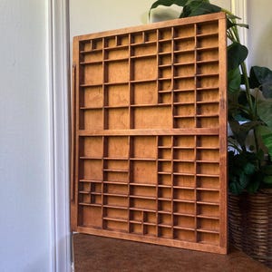 May include: A vintage wooden printer's tray with numerous small compartments of varying sizes. The tray is made of light brown wood and is designed to hold small items. It is displayed on a brown table with a gold trim, with a green plant in the background.