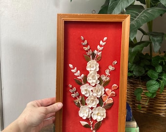 May include: A framed floral arrangement with white shell flowers and brown leaves against a red background. The wooden frame has a warm, golden hue. The artwork is a decorative piece.