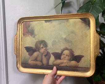 May include: A rectangular serving tray with a textured gold-painted frame. The center features a printed image of two cherubs with wings resting their arms on a ledge against a cloudy, pale yellow background, inspired by classical Renaissance art.