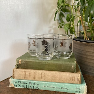 May include: Four frosted glass tumblers with silver leaf designs of leaves. The glasses are stacked on top of three vintage books. The top book is green, the middle book is tan, and the bottom book is blue.