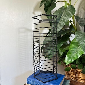 May include: A tall, rectangular black metal wire storage rack with multiple horizontal slots. The rack features a sturdy frame with mounting holes at the top and bottom, designed for organizing media or small items in a vertical orientation.