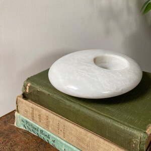 May include: White marble candle holder with a round, slightly concave top and a circular opening in the centre. The holder is sitting on top of a stack of three books.