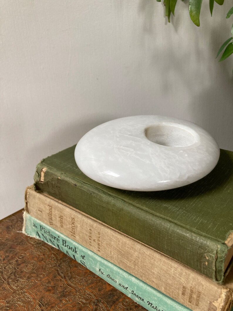 May include: White marble candle holder with a round, slightly concave top and a circular opening in the centre. The holder is sitting on top of a stack of three books.