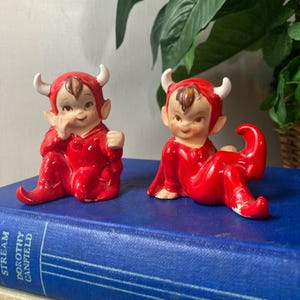 May include: Two small, vintage devil figurines. Each figurine is red with white horns, brown hair, and a smiling face. One is seated, the other is crawling. They are displayed on a stack of blue and green books.