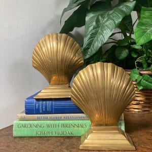 May include: Two gold-colored seashell bookends. The bookends are placed on top of a stack of books with titles like "The Deepening Stream" and "Gardening with Perennials". The background includes green plants and a wooden surface.