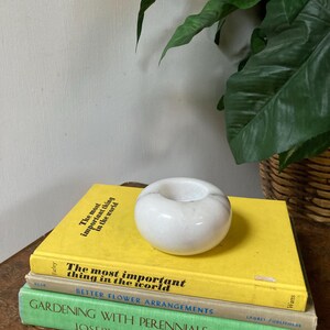 May include: A stack of four books with yellow, beige, green, and brown covers. The top book has the text "The most important thing in the world" on the cover. A white marble candle holder sits on top of the books.