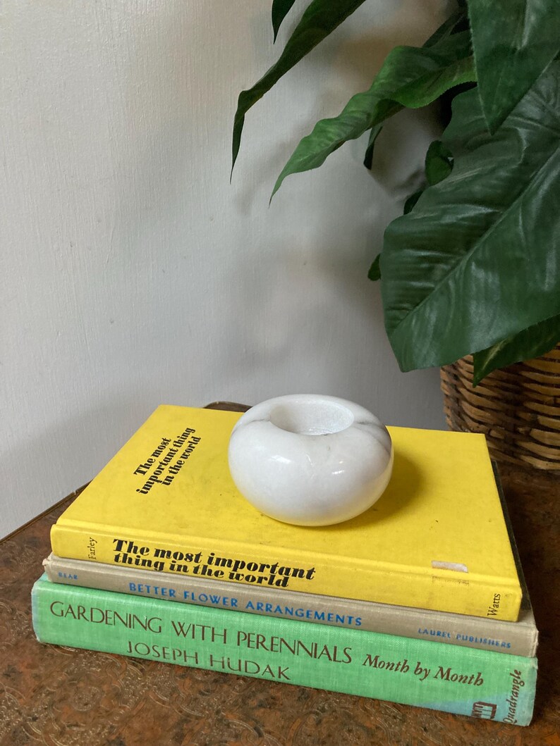 May include: A stack of four books with yellow, beige, green, and brown covers. The top book has the text "The most important thing in the world" on the cover. A white marble candle holder sits on top of the books.
