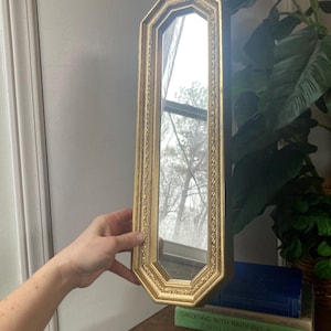 May include: A gold-framed, octagonal mirror with ornate detailing. The mirror is held up against a wall, reflecting a window and the surrounding environment. The frame has a vintage aesthetic.