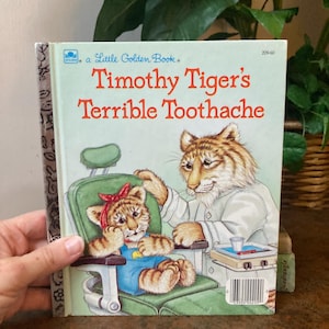 May include: A Little Golden Book titled "Timothy Tiger's Terrible Toothache." The cover features an illustration of a tiger cub in a dentist's chair, with a concerned expression. The dentist, also a tiger, is examining the cub's mouth.