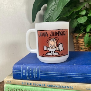 May include: White coffee mug with a handle, featuring a cartoon character and the words "JAVA JUNKIE!" on a red background. The mug is sitting on a stack of books with blue and green covers.
