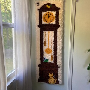 May include: A long, rectangular wall hanging featuring a brown grandfather clock design. The clock face is yellow with black dots, and the pendulum and weights are visible. A yellow cat sits at the base. The background is off-white with a shaggy texture.