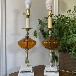 May include: A pair of amber glass table lamps with white marble bases and gold accents. The lamps have a swirl design and are lit with white light bulbs.