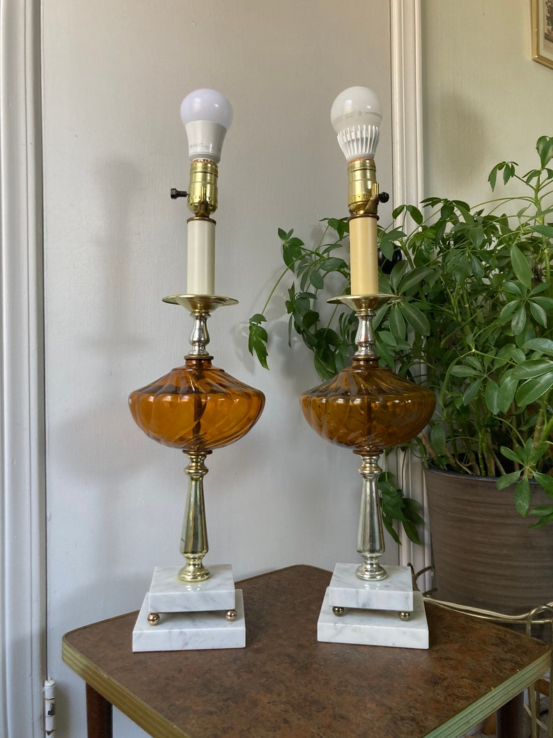 May include: A pair of amber glass table lamps with white marble bases and gold accents. The lamps have a swirl design and are lit with white light bulbs.