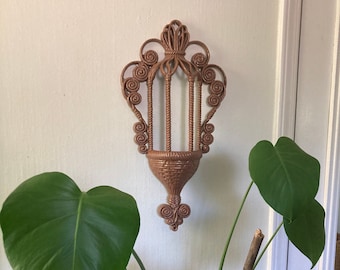 May include: A brown wicker wall hanging planter with a decorative design. The planter has a basket-like shape with a rounded top and a pointed bottom. The planter is made of woven wicker and has a rustic look.