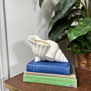 May include: A white ceramic seashell-shaped planter resting on a stack of three books. The books are titled The Deepening Stream by Dorothy Canfield, Better Flower Arrangements, and Gardening with Perennials Month by Month by Joseph Hudak.