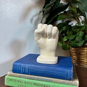 May include: A glossy, off-white ceramic sculpture shaped like a clenched fist, resting on a rectangular base. The sculpture is positioned atop a stack of three hardcover books, including titles like The Deepening Stream and Gardening with Perennials.