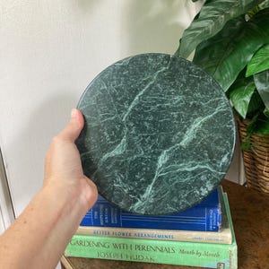 May include: A circular dark green marble slab with white veining patterns throughout the stone surface. The polished stone disc features a smooth texture and natural mineral variations, showcasing a deep forest green color palette with light streaks.