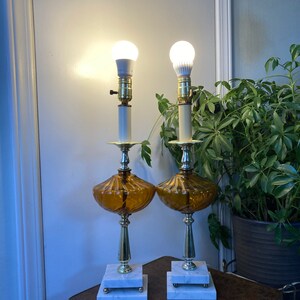 May include: A pair of amber glass table lamps with white marble bases and gold accents. The lamps have a ribbed design and are lit with white light bulbs.