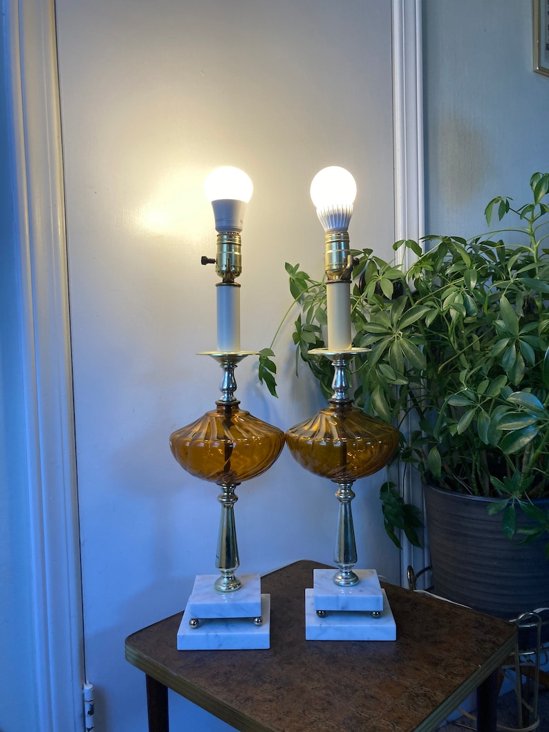 May include: A pair of amber glass table lamps with white marble bases and gold accents. The lamps have a ribbed design and are lit with white light bulbs.