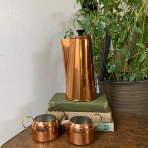 May include: A copper coffee pot with a black handle and a matching set of two copper creamer and sugar bowls with gold handles. The coffee pot is sitting on a stack of books.