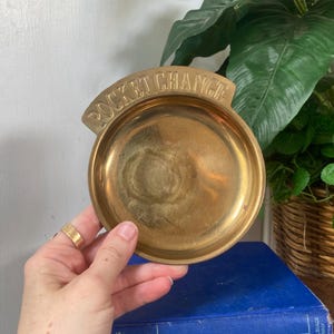 May include: A vintage brass pocket change dish with the words "POCKET CHANGE" embossed on the top. The dish is round with a shallow bowl and a curved top. The dish is held by a hand.