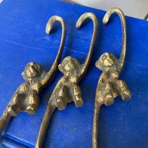 May include: Three brass monkey-shaped hooks. Each hook features a monkey figure with its arms raised, forming the hook. The hooks are a warm, antique gold color and are set against a blue background.