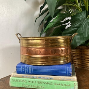 May include: A round brass and copper planter with two small side handles. The exterior features horizontal ribbed detailing and a central band of embossed circular patterns. The metal has a polished finish with visible warm gold and copper tones.