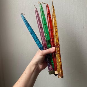 May include: A hand holds five colorful taper candles. The candles are blue, purple, green, red, and yellow. Each candle is filled with glitter and has a metallic base. The candles are approximately 10 inches long.