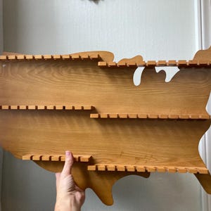 May include: Wooden shelf shaped like the United States of America with three shelves. Each shelf has slots for displaying items.