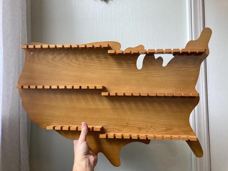 May include: Wooden shelf shaped like the United States of America with three shelves. Each shelf has slots for displaying items.