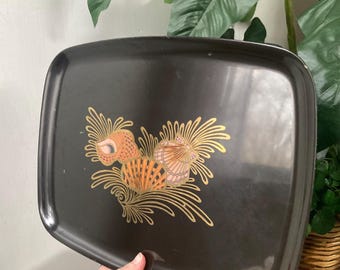 May include: A black rectangular serving tray with rounded corners. The tray features a seashell design in shades of orange, pink, and gold. The tray is held by a person's hand.