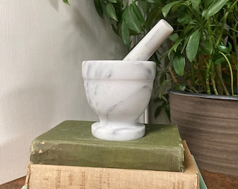 May include: A white marble mortar and pestle set rests atop a stack of vintage books. The books are in shades of green, tan, and blue, with visible text. A leafy green plant is in the background.