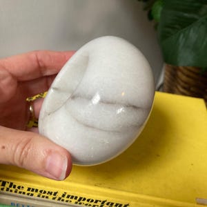 May include: White marble ring holder with a smooth, rounded surface and subtle gray veining. The holder is shaped like a donut with a hole in the center.
