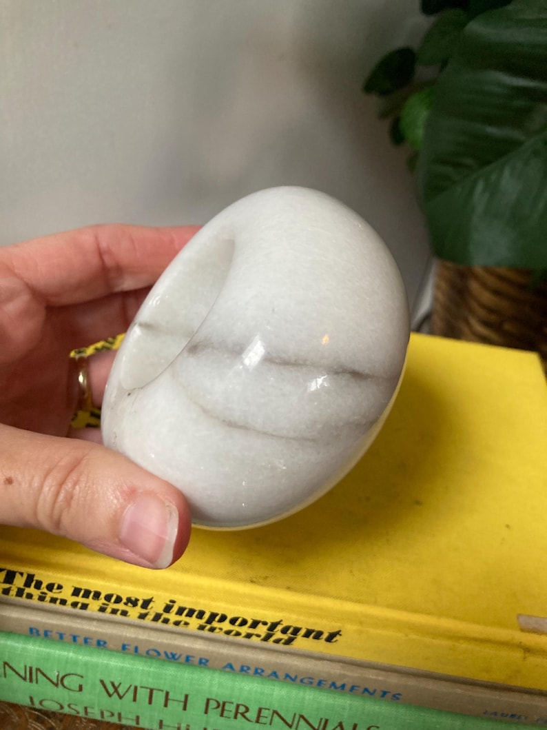 May include: White marble ring holder with a smooth, rounded surface and subtle gray veining. The holder is shaped like a donut with a hole in the center.
