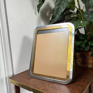 May include: A rectangular picture frame featuring a dual-tone design with a polished silver outer border and a gold inner border. The frame has rounded corners and contains a plain, light brown rectangular insert within the center area.