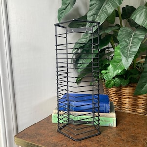 May include: A black metal CD rack with a rectangular shape. The rack is made of thin, vertical bars and sits on a brown and gold table. A stack of books is visible behind the rack, and a large green plant is in the background.