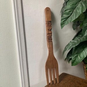 May include: A large, wooden fork with a carved handle and four tines. The handle features detailed carvings. The fork is a light brown color and appears to be handmade. It is leaning against a white wall and a brown table.