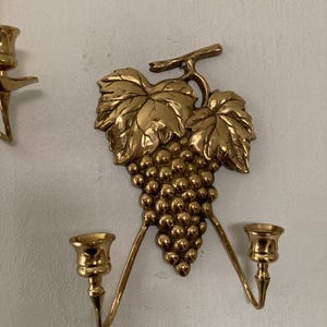 May include: Gold-colored wall sconce featuring a grape cluster and leaves design. The sconce has two candle holders extending outwards. The decorative piece is made of metal and is mounted on a white wall.