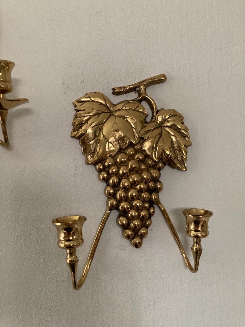 May include: Gold-colored wall sconce featuring a grape cluster and leaves design. The sconce has two candle holders extending outwards. The decorative piece is made of metal and is mounted on a white wall.