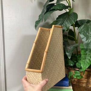 May include: A woven magazine holder with a bamboo frame. The holder is made of a light tan woven material and has a rectangular shape. The bamboo frame is a light brown color. The holder is sitting on top of a stack of books.