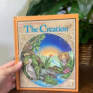 May include: A hardcover book titled A Golden Bible Story The Creation with an orange spine and border. The cover features a circular illustration of a landscape with a tiger, alligator, bird, and sun, set against a gold-toned background.