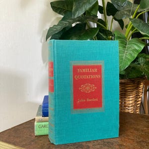 May include: A teal hardcover book titled "Familiar Quotations" by John Bartlett. The book has a red rectangular label with gold lettering and decorative elements. Other books are stacked behind it on a wooden table.
